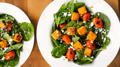 Two plates of spinach salad with roasted orange squash and cherry tomatoes on a wooden table