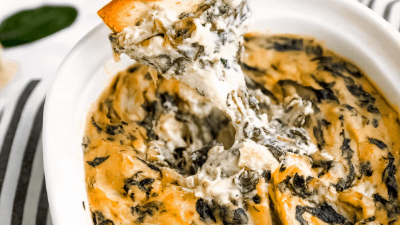 Bread slice dipped in creamy spinach artichoke dip in a white bowl