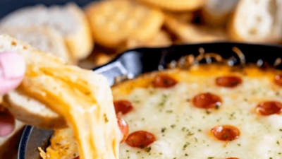 Cheesy baked pasta with pepperoni slices, served in a black pan as a hand lifts a cheesy piece