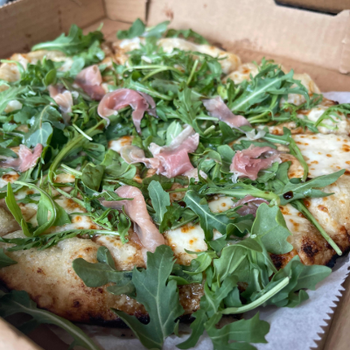 Pizza topped with arugula and prosciutto in a cardboard box