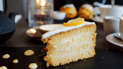 Slice of lemon cake with white frosting on a dark table, with coffee cups and pastries blurred in the background