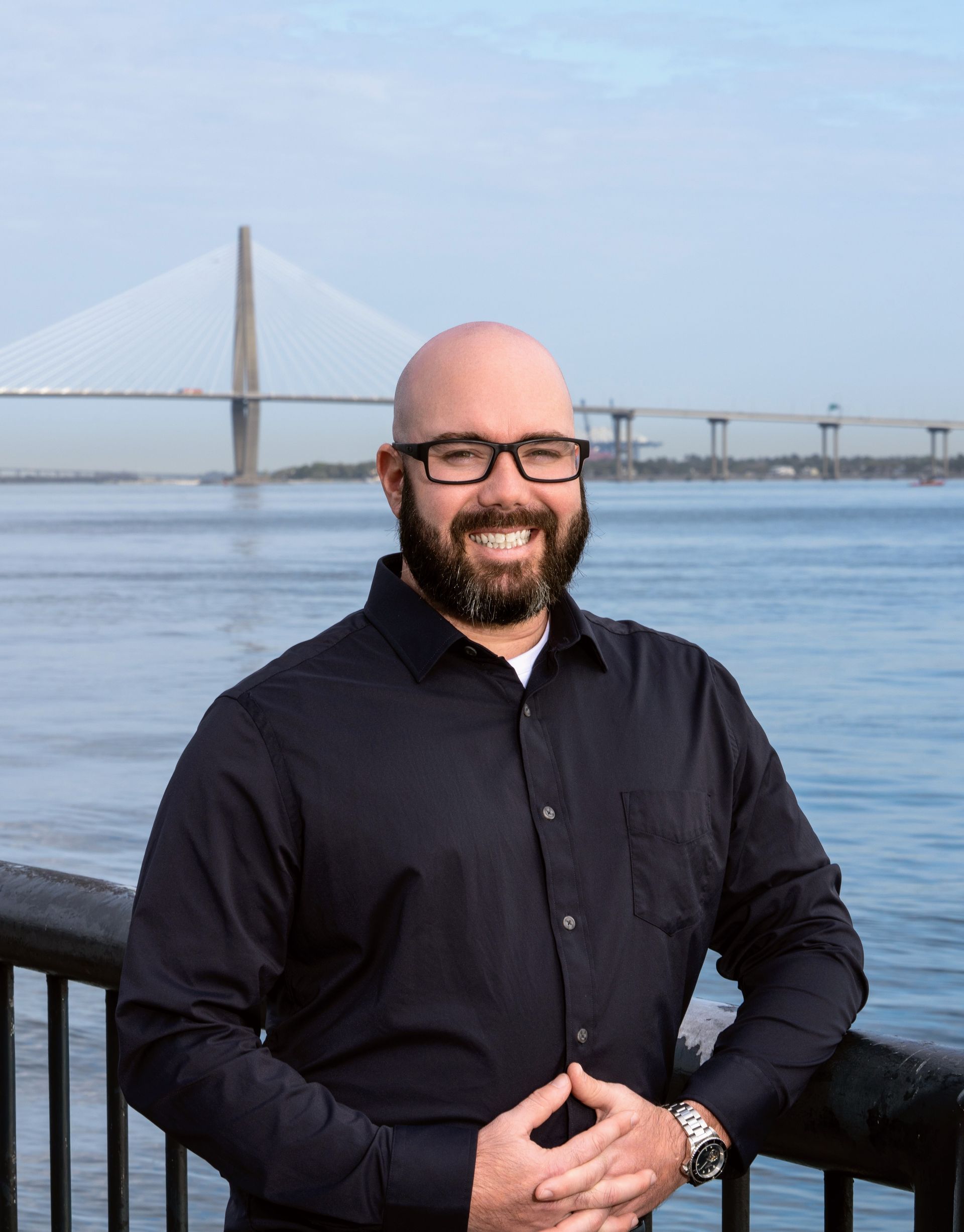 Smiling man in a black shirt standing by a waterfront railing with a bridge in the background