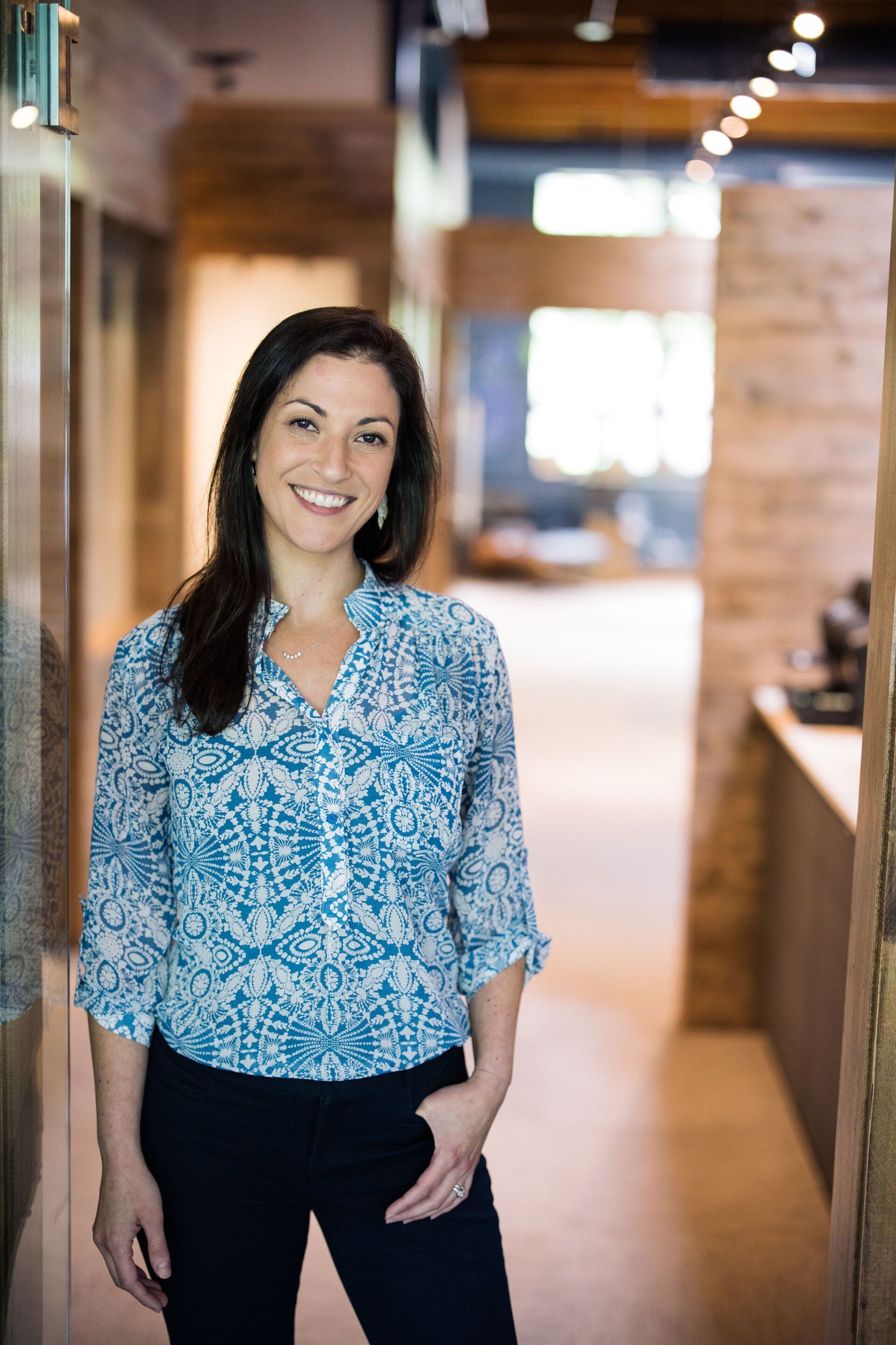 Woman smiling in a blue patterned blouse, standing in a warmly lit office hallway.