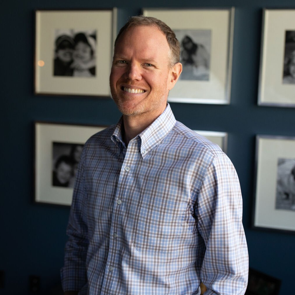 Smiling man in a blue checkered shirt standing in front of framed photos on a wall