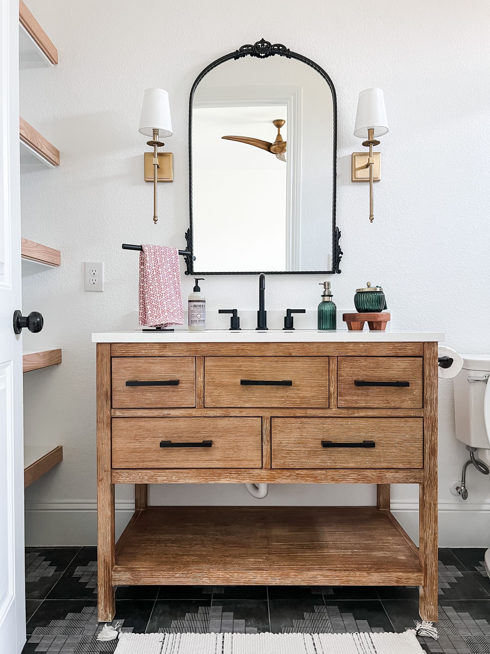 Bathroom with wooden vanity, ornate mirror, gold sconces, and patterned floor.