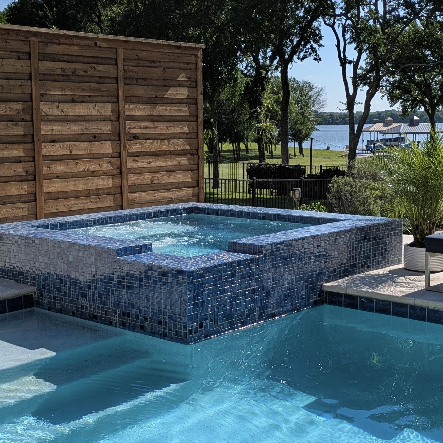 Blue-tiled hot tub and pool overlook a lake, bordered by a wooden privacy fence, trees, and greenery.