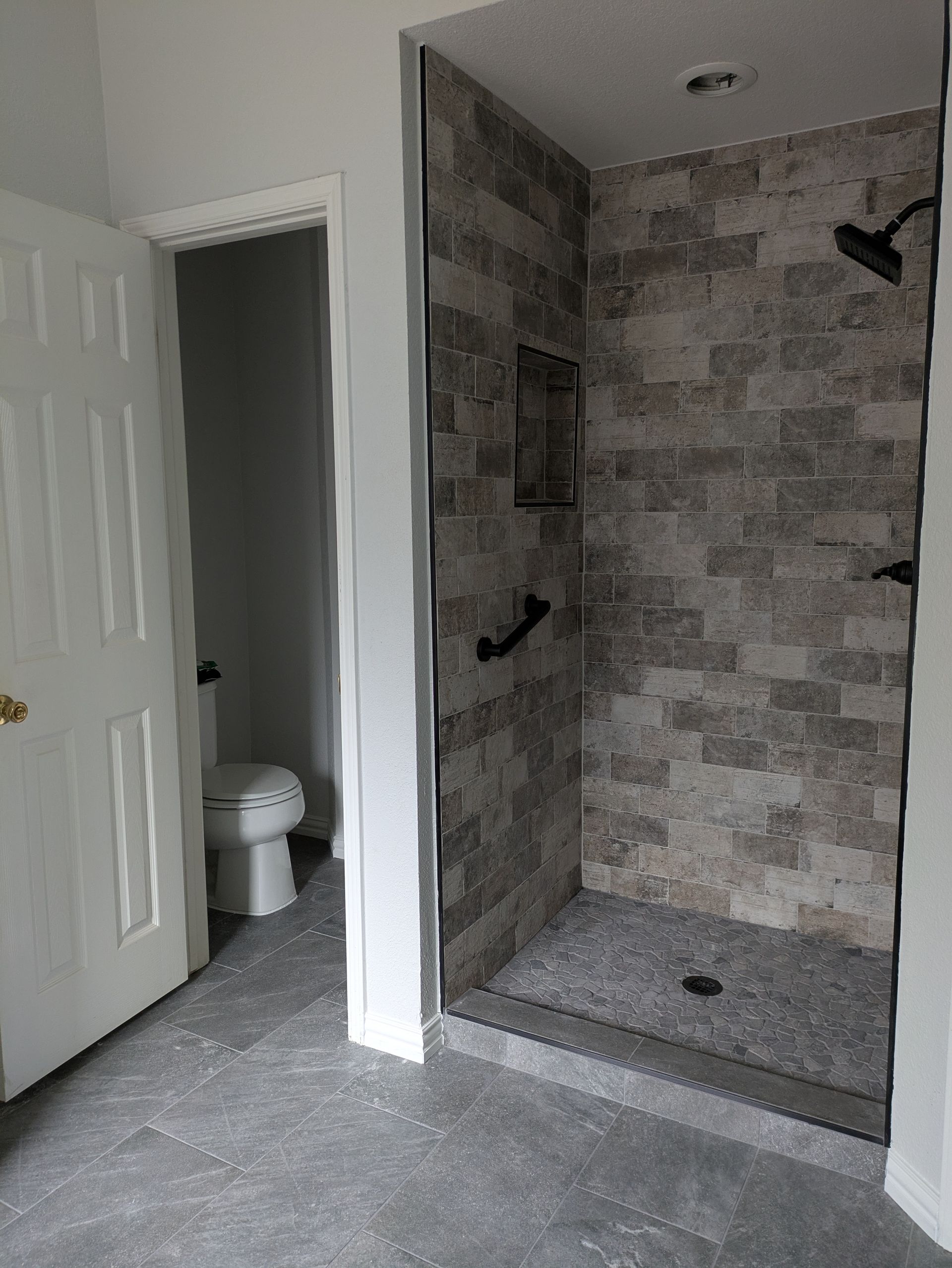 Bathroom with a tiled shower, toilet, and open door. Gray and neutral tones throughout.