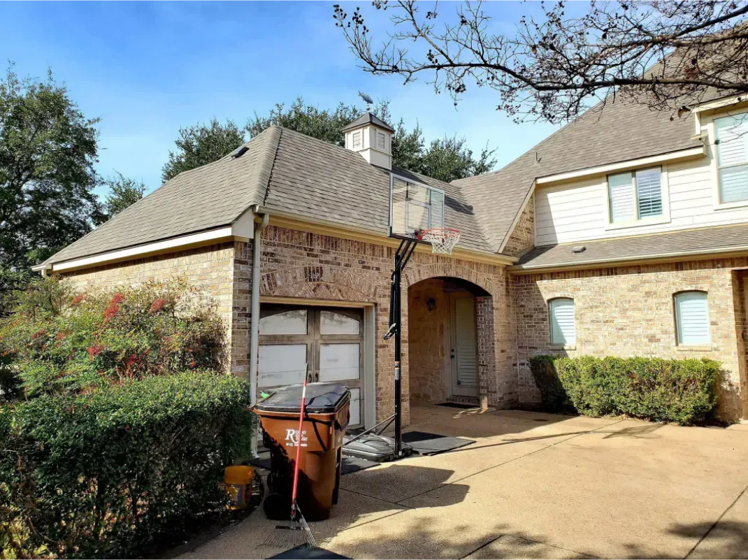 Beige brick house with attached garage and driveway; trash can and shrubs in front.