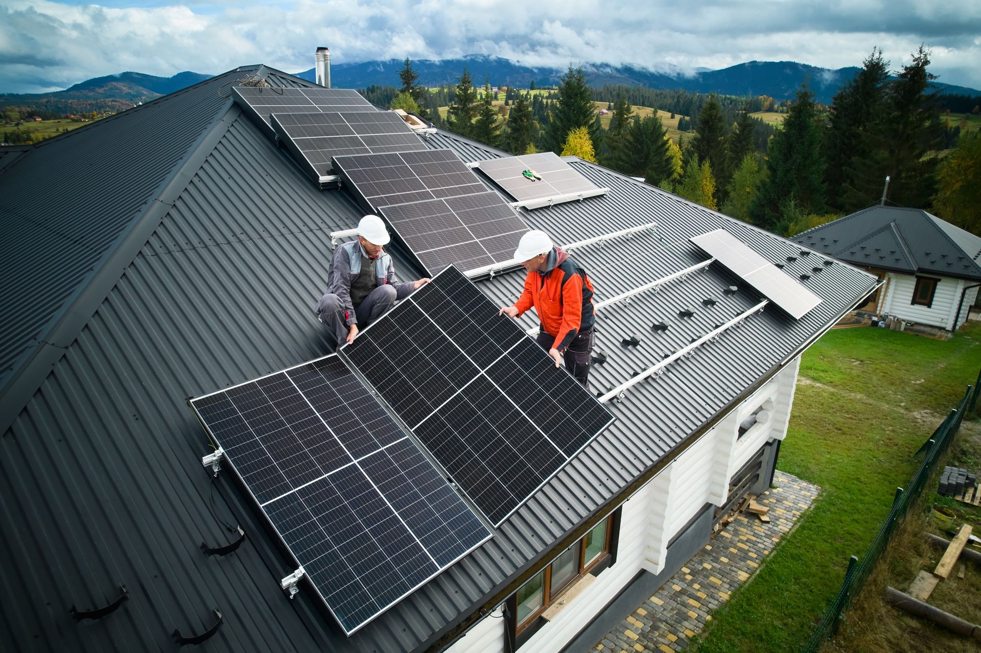 Two workers installing solar panels on a house roof. Blue sky, mountains in the background.