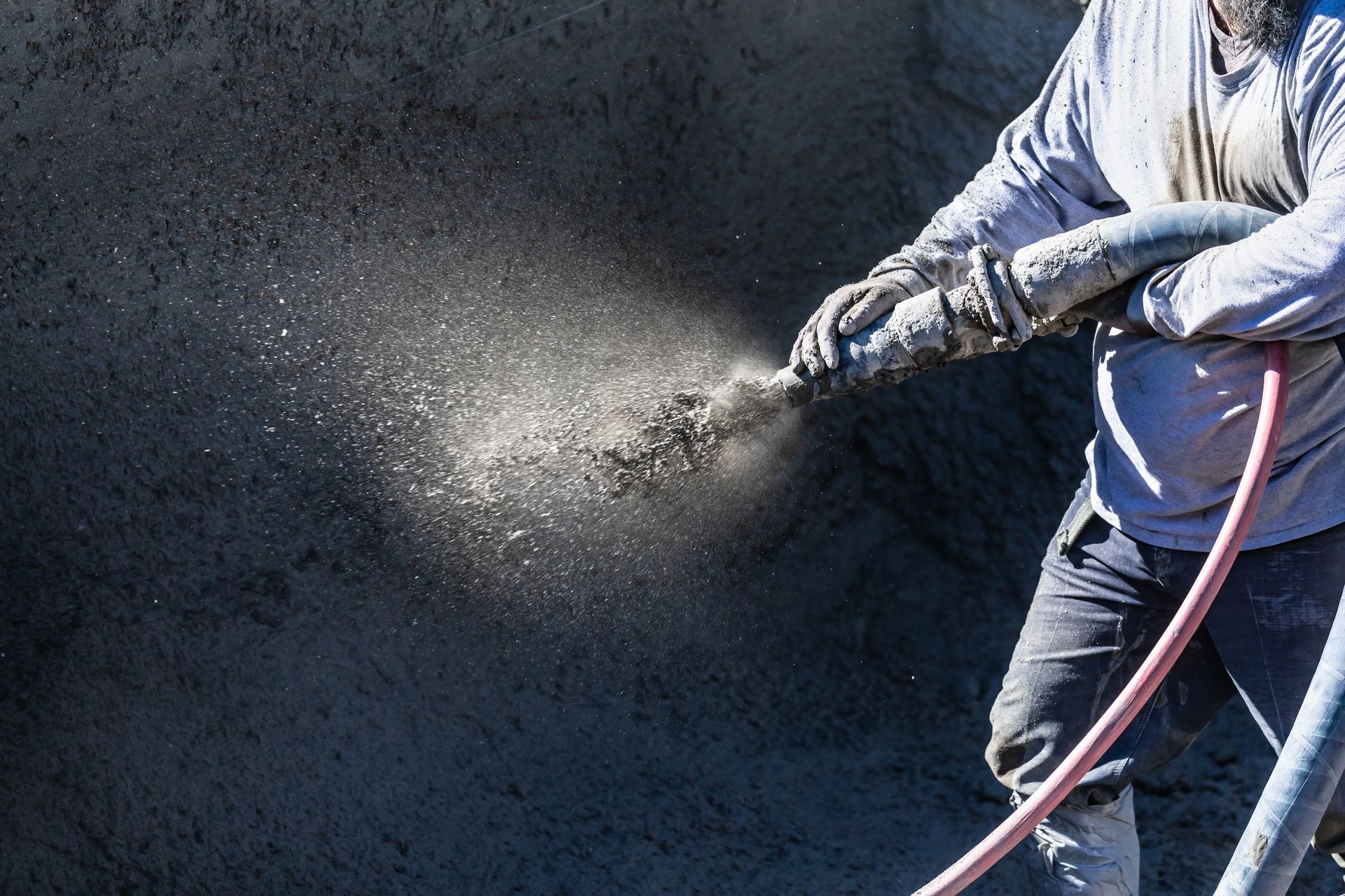Worker spray-painting a dark wall, creating a fine mist through a hose-connected spray gun