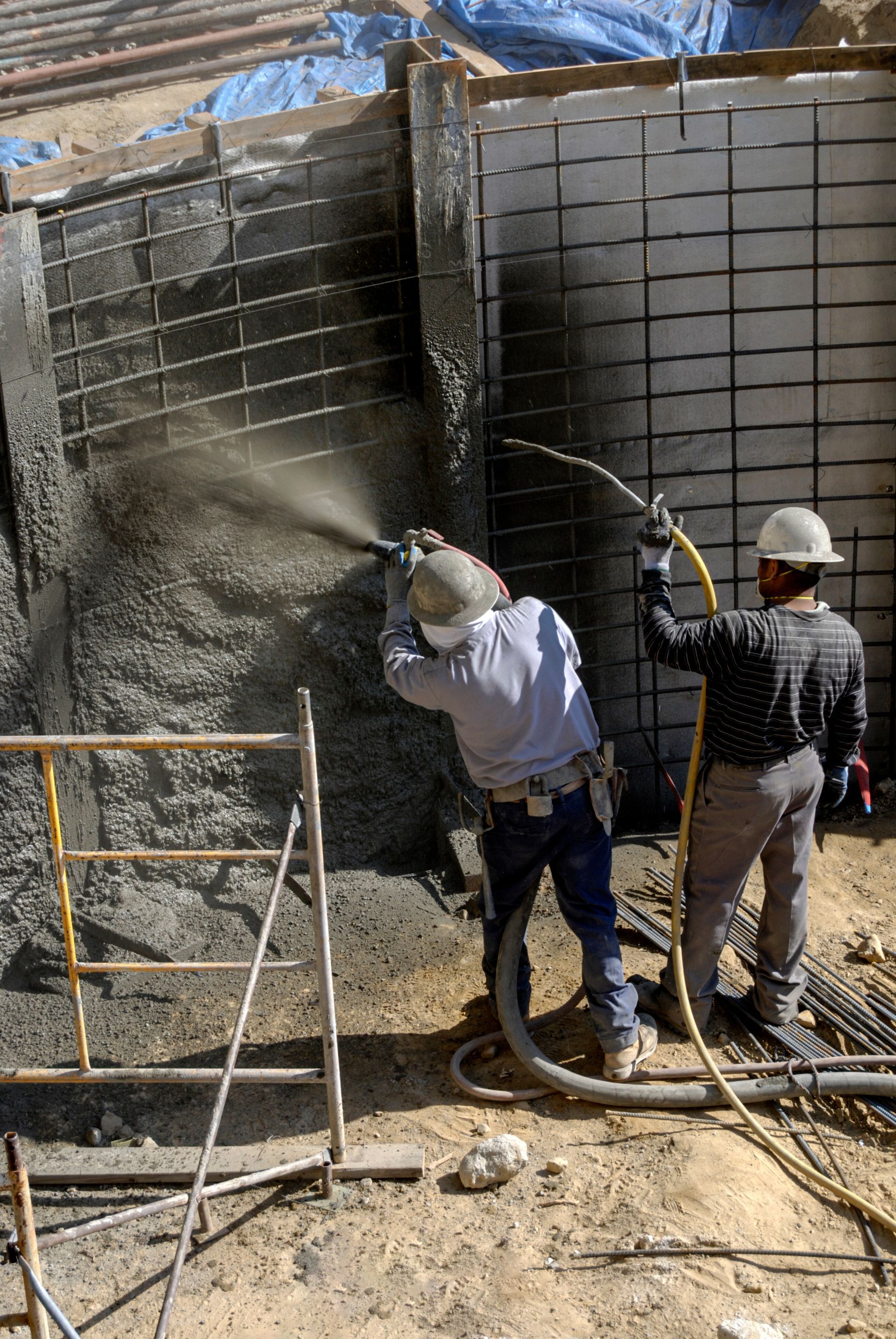 Workers spray concrete onto a wall reinforcement grid at a construction site. Workers spray concrete onto a wall reinforcement grid at a construction site.