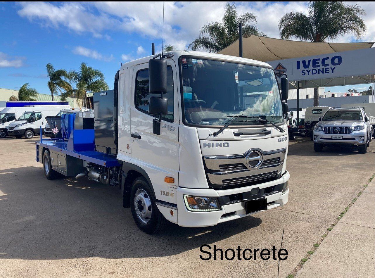 White Iveco flatbed truck at a dealership, with blue equipment on the back and “Shotcrete” text visible.