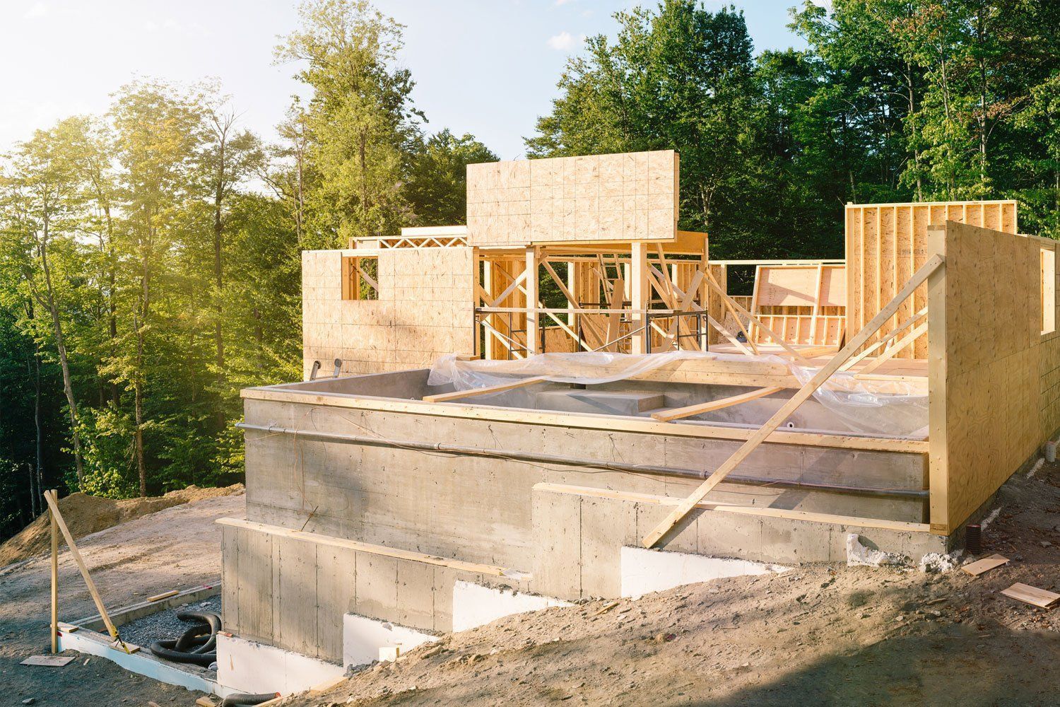 House under construction with unfinished wood framing and concrete foundation surrounded by trees House under construction with unfinished wood framing and concrete foundation surrounded by trees