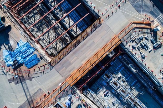 Aerial view of a construction site with steel framework, blue tarps, and orange scaffolding around a concrete roadway.