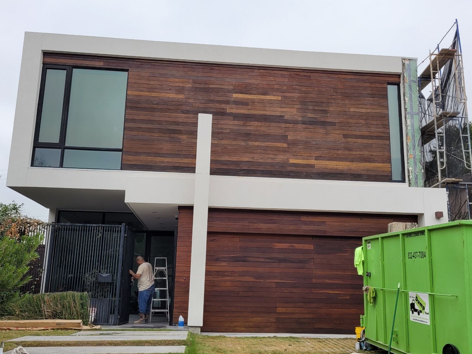 A man is standing in front of a modern house with a green dumpster in front of it.