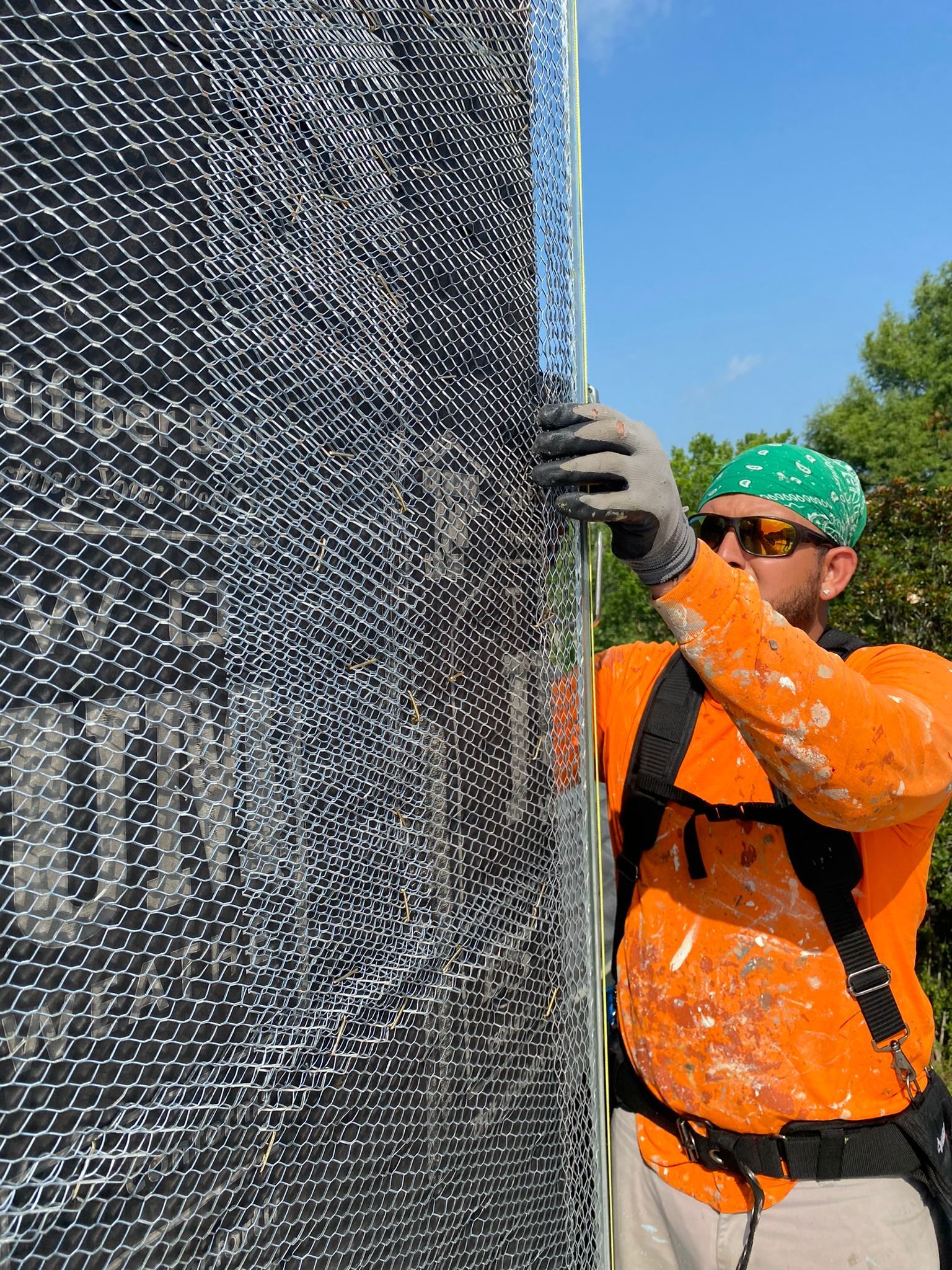 A man in an orange shirt and green bandana is standing next to a wall.