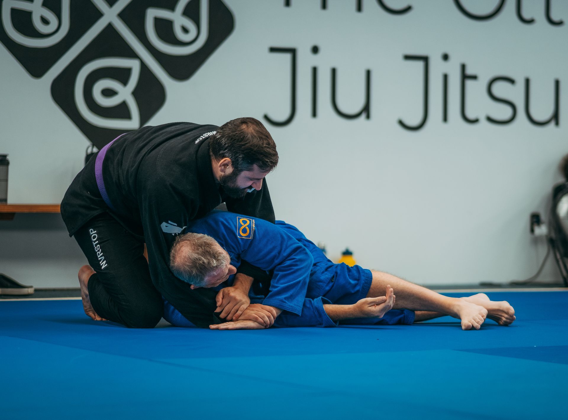 Two jiu-jitsu practitioners grapple on blue mats in the best martial arts center in Ottawa, ON, with a wall sign in the background.