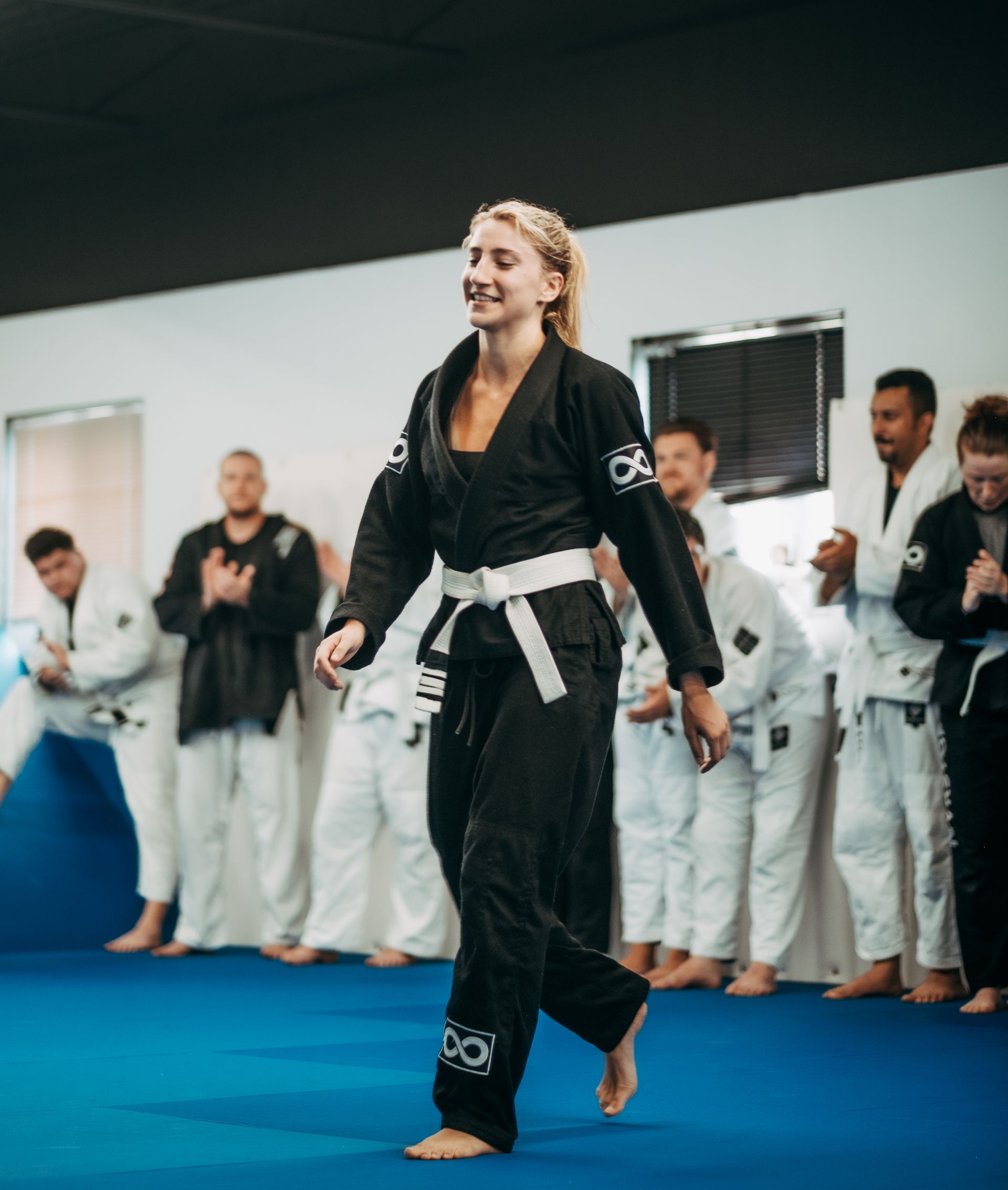 Martial arts class with a person in a black gi and white belt walking on blue mats, others watching behind at the best martial arts center in Ottawa, ON
