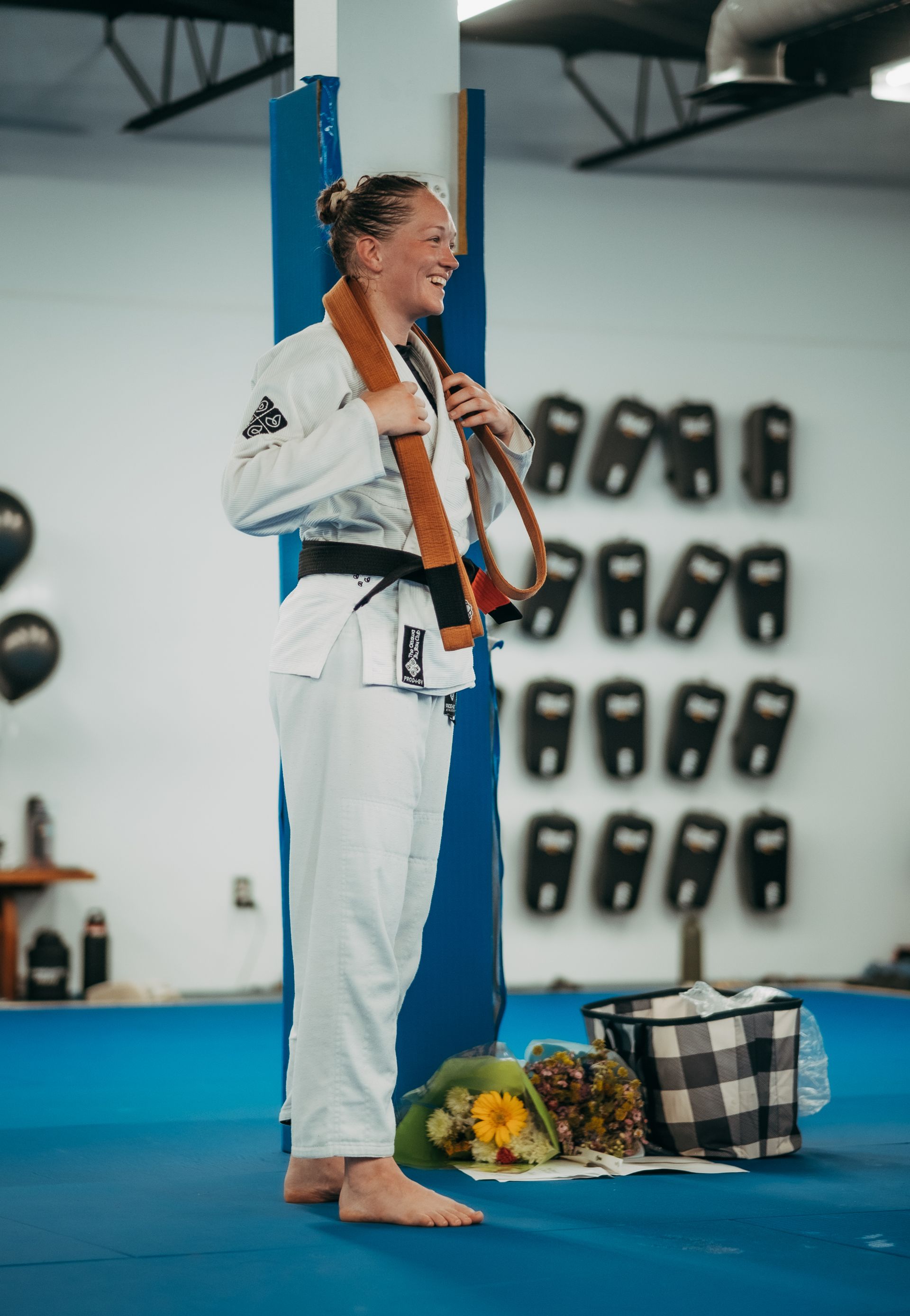 Person in a white martial arts gi holding a medal, standing on a blue mat with flowers and a basket nearby at the best martial arts center in Ottawa, ON