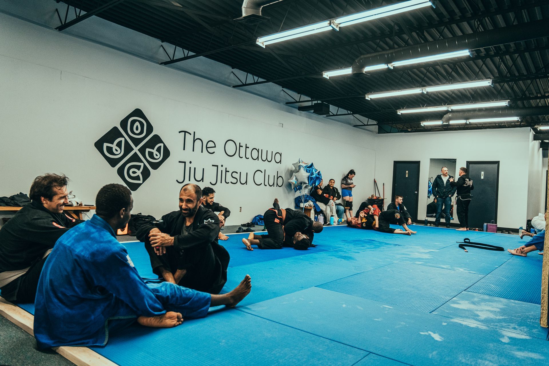 Martial arts class on blue mats in a dojo, with students seated and practicing near a wall logo at the best martial arts center in Ottawa, ON