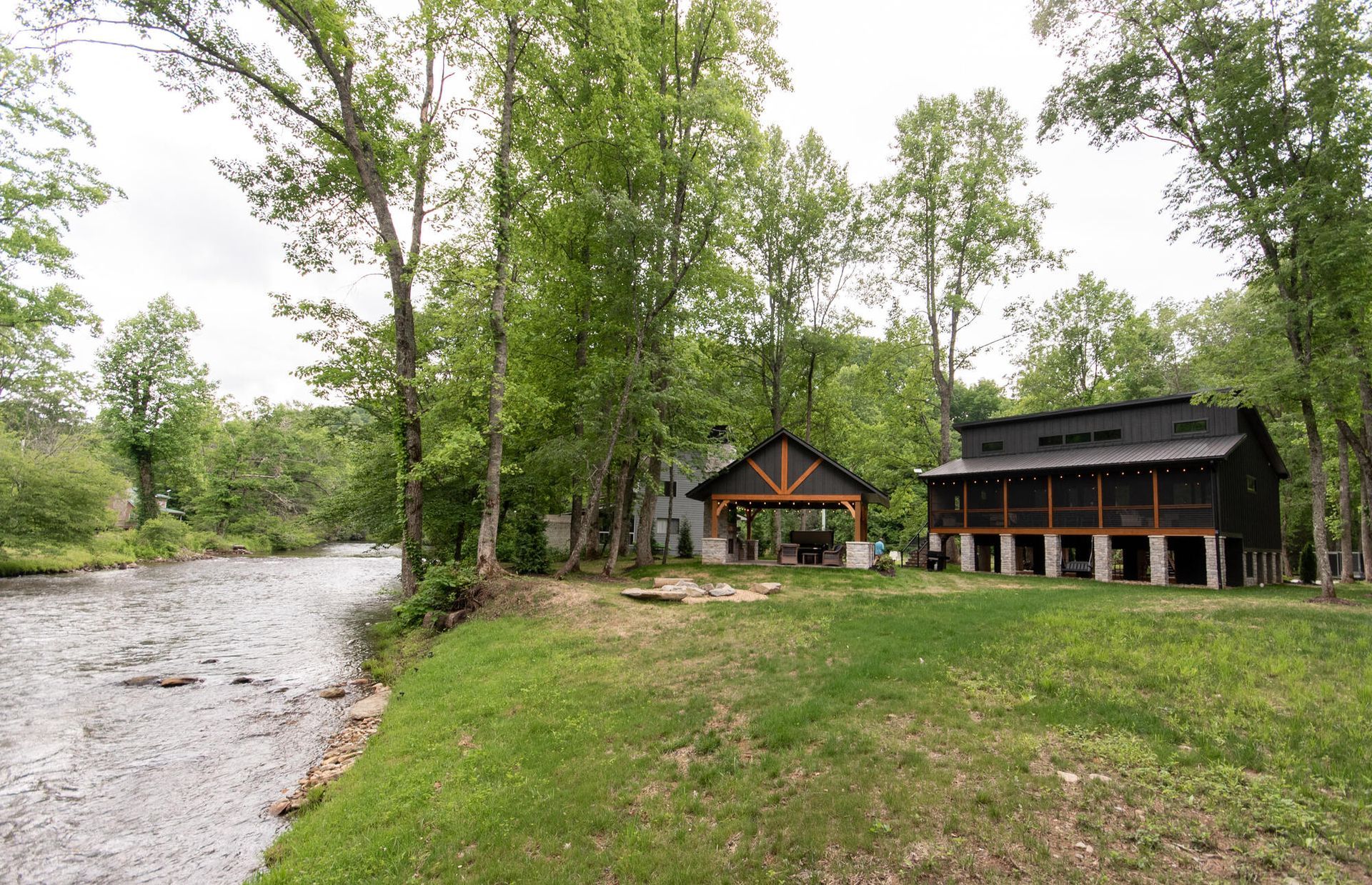 Riverside cabin and lodge beside a grassy clearing and trees under a cloudy sky