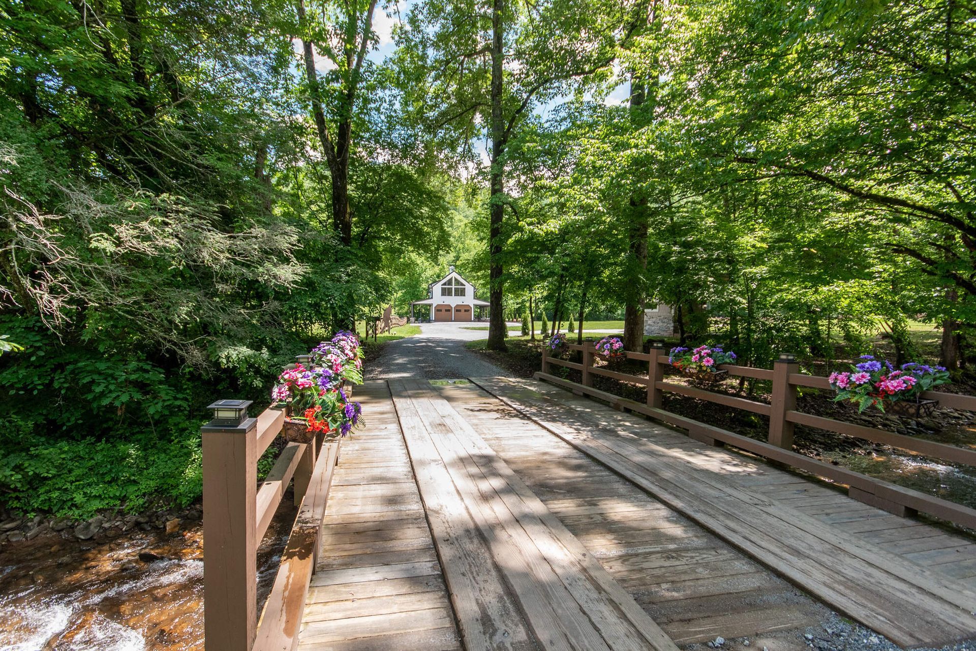 Wooden walkway through a green wooded park, lined with flowers, leading to a small white pavilion.
