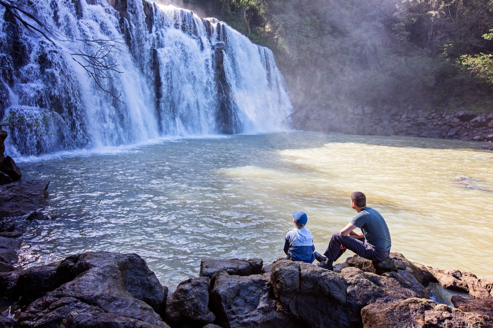 A man and a child are sitting on rocks near a waterfall.