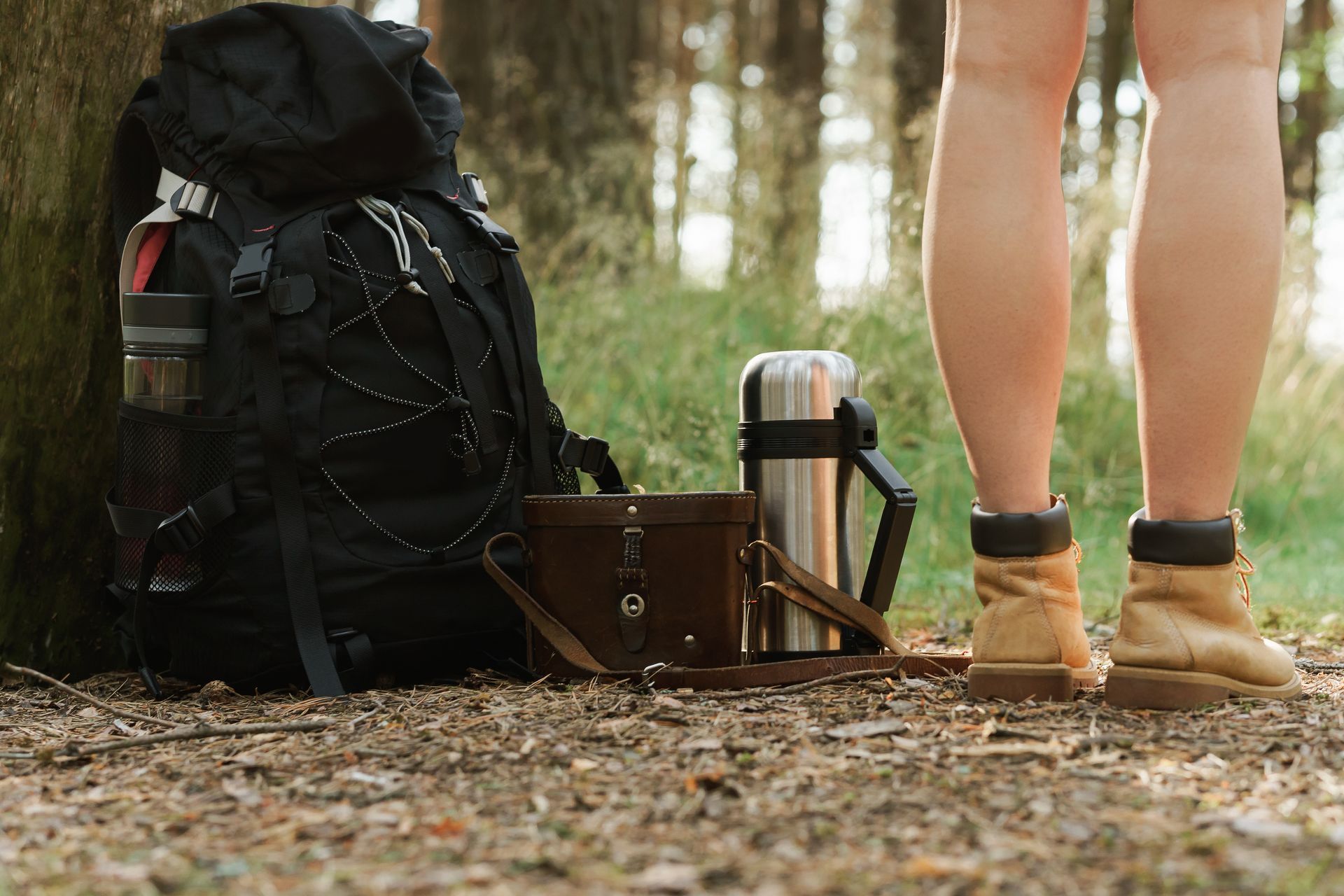 A person is standing next to a backpack and a thermos in the woods.