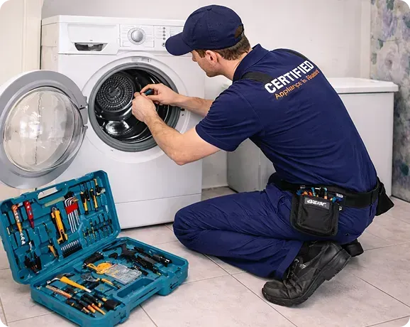 Worker in a navy uniform kneeling at an open front-load washer beside a toolbox