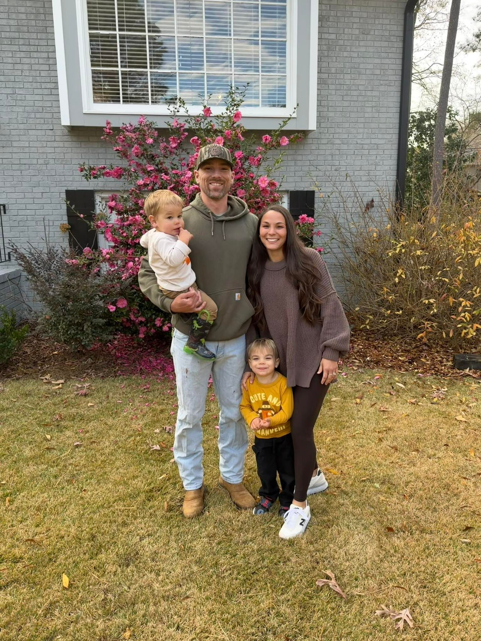 Family of four posing on a lawn in front of a house, with a flowering bush behind them.