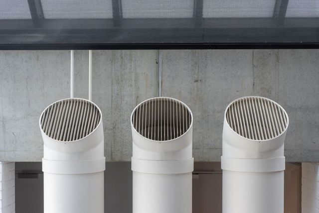 Three white rooftop vent pipes with metal mesh caps against a concrete wall.