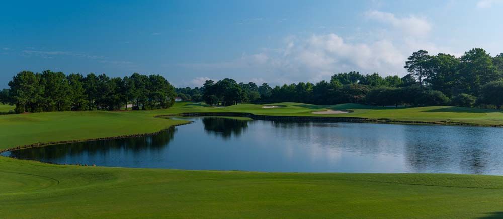 A serene golf course scene featuring a lake reflecting the sky and surrounding greenery, under a clear blue sky.