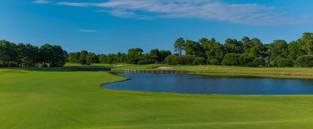 Green golf course with a body of water and trees under a blue sky. A bridge crosses the water.