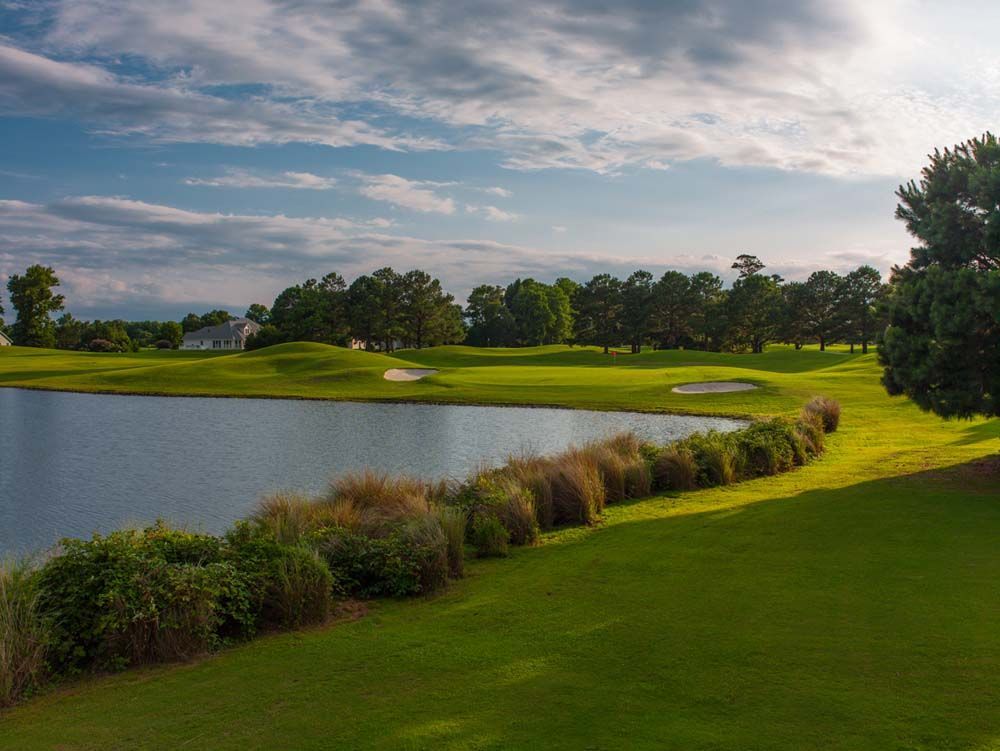 A lush golf course with a pond and green grass, under a cloudy blue sky.