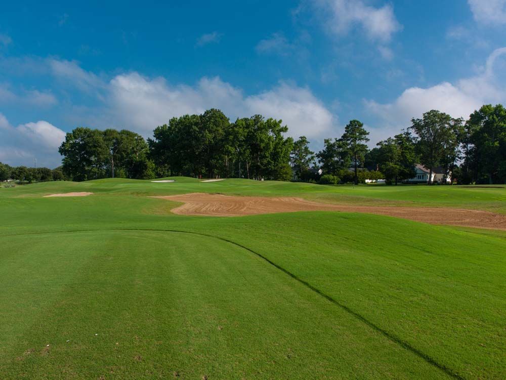 Green golf course with lush grass, a brown patch, and trees against a blue sky with scattered clouds.