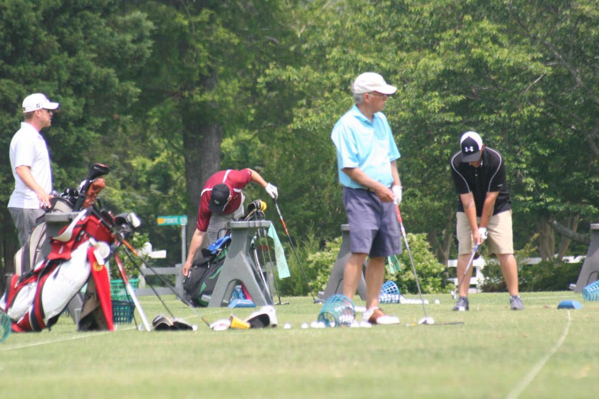 Several golfers on a grassy driving range. One person watches as another prepares to swing, while others stand near golf bags.