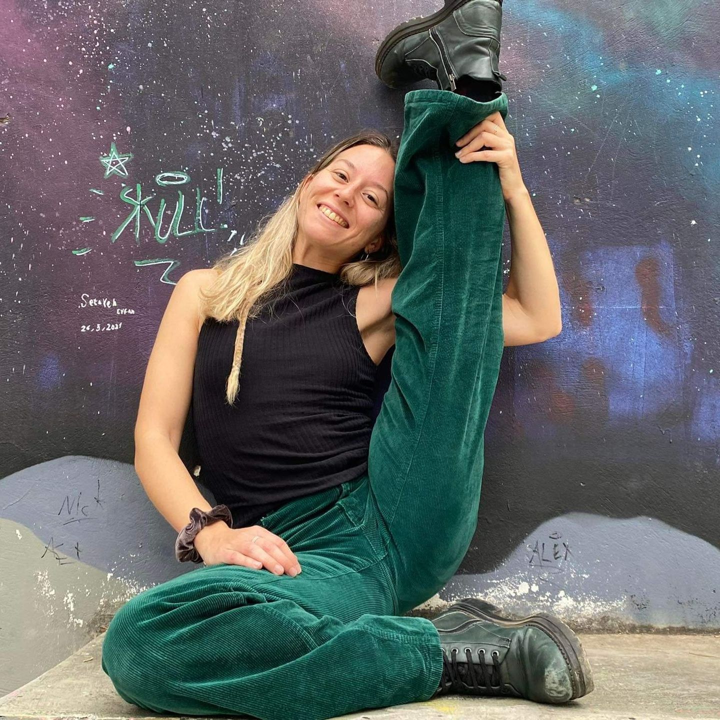 Woman in green pants and black top does the splits against a graffiti wall, smiling.