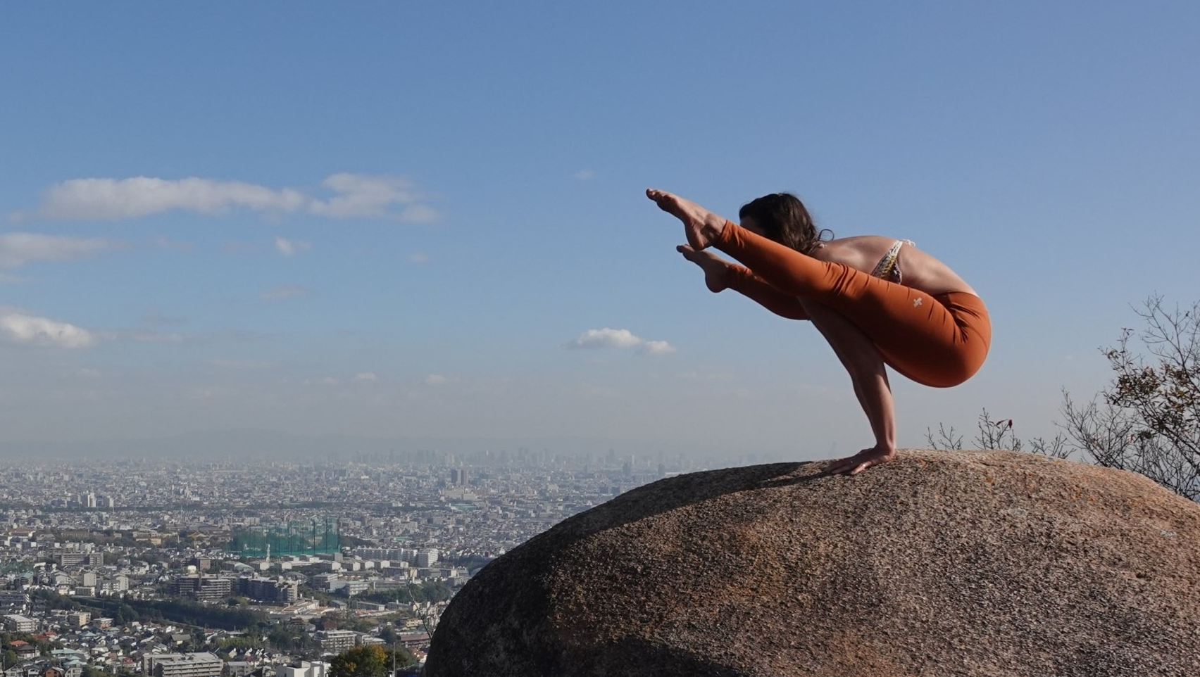 Person balances in a yoga pose atop a large rock, overlooking a cityscape. Blue sky.