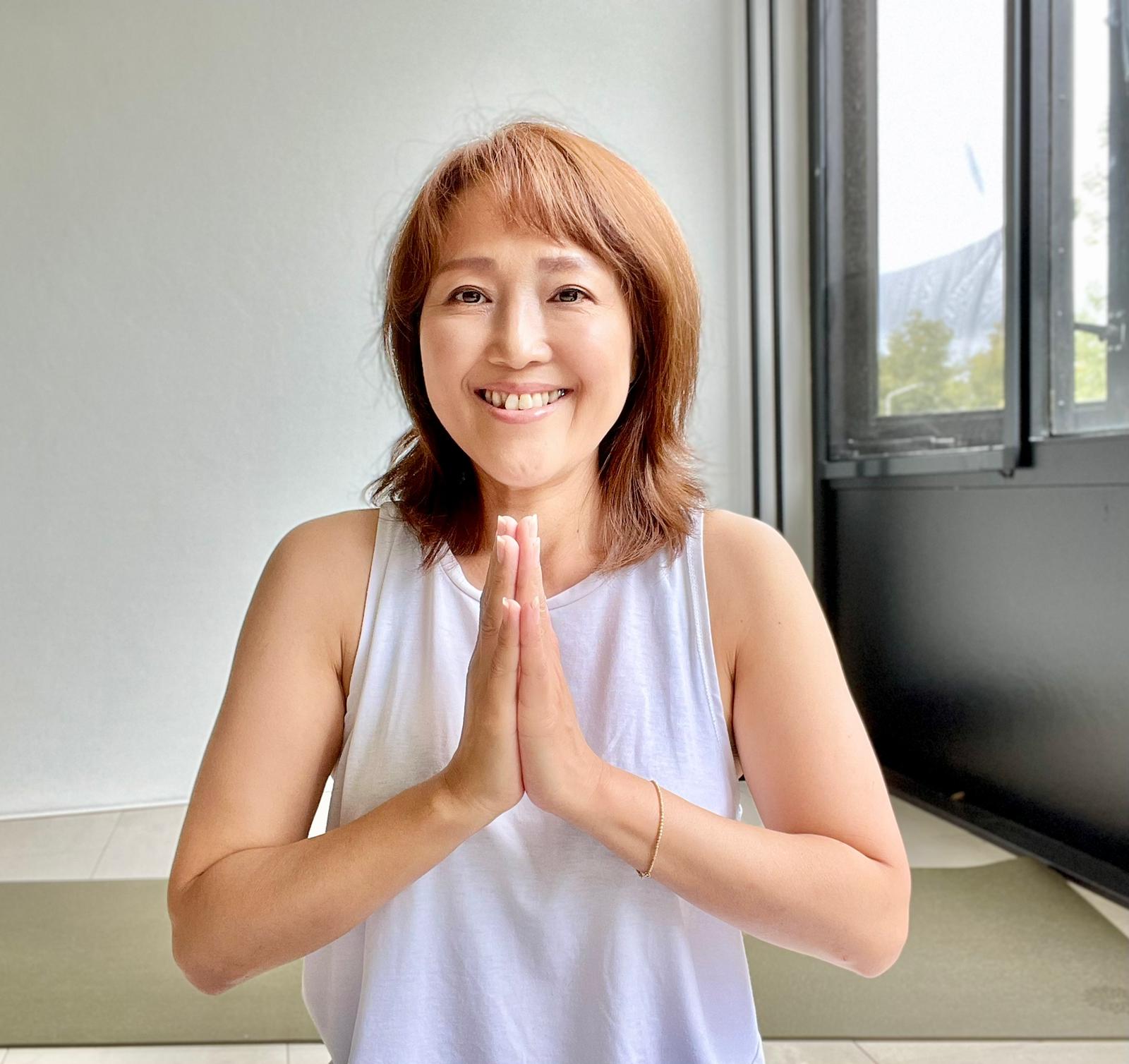Woman in white tank top, hands in prayer pose, smiling indoors.