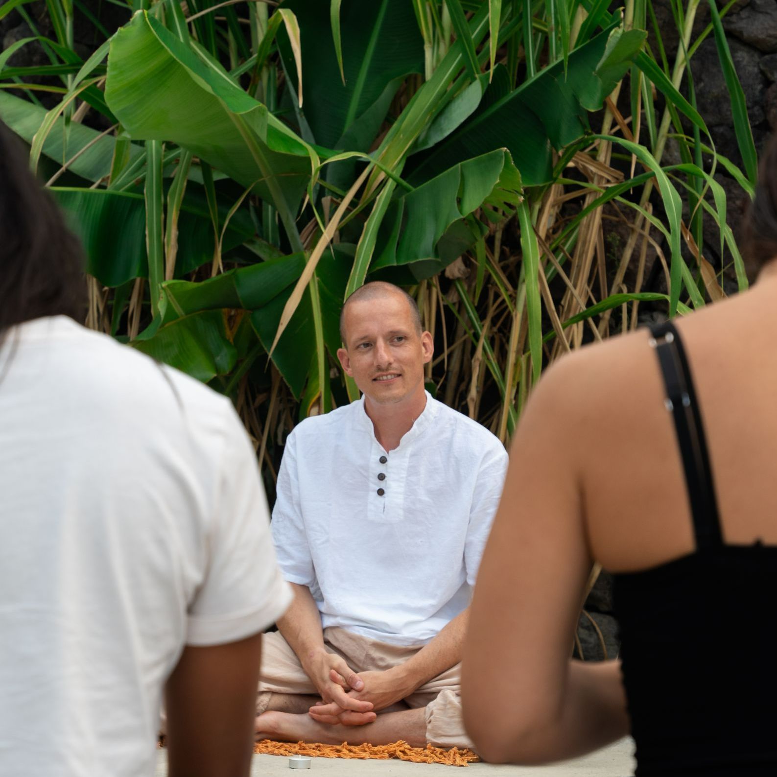 Man in white shirt and beige pants sits cross-legged, facing two people outdoors.
