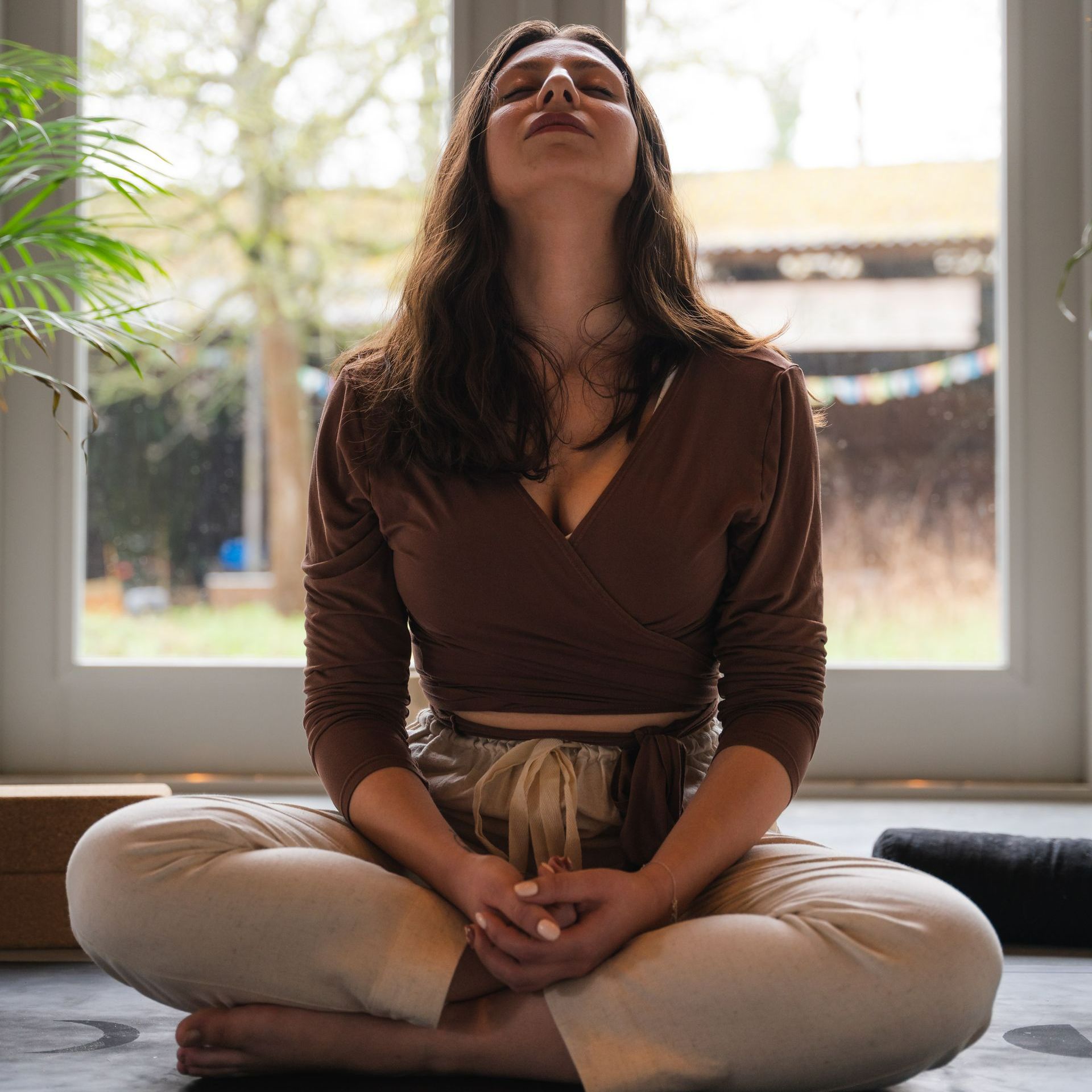 Woman in brown top and beige pants meditating with eyes closed in a bright room.