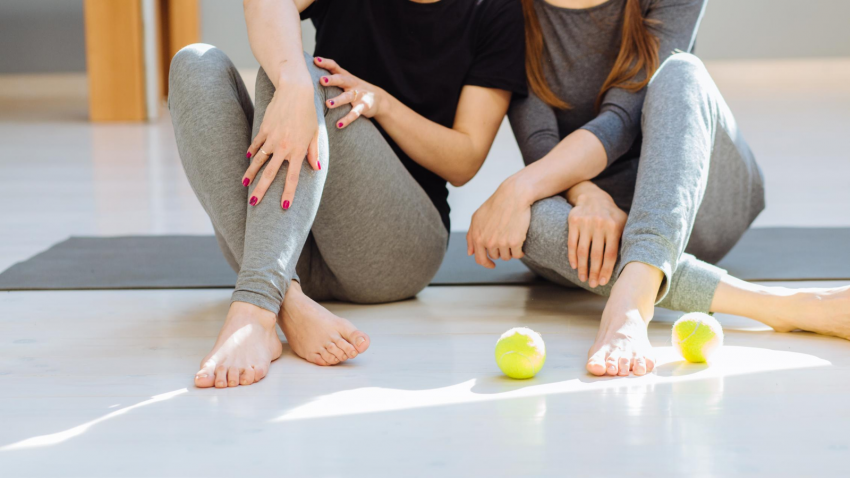 Two people in gray workout pants sit on a floor, touching. Two green apples are on the floor.
