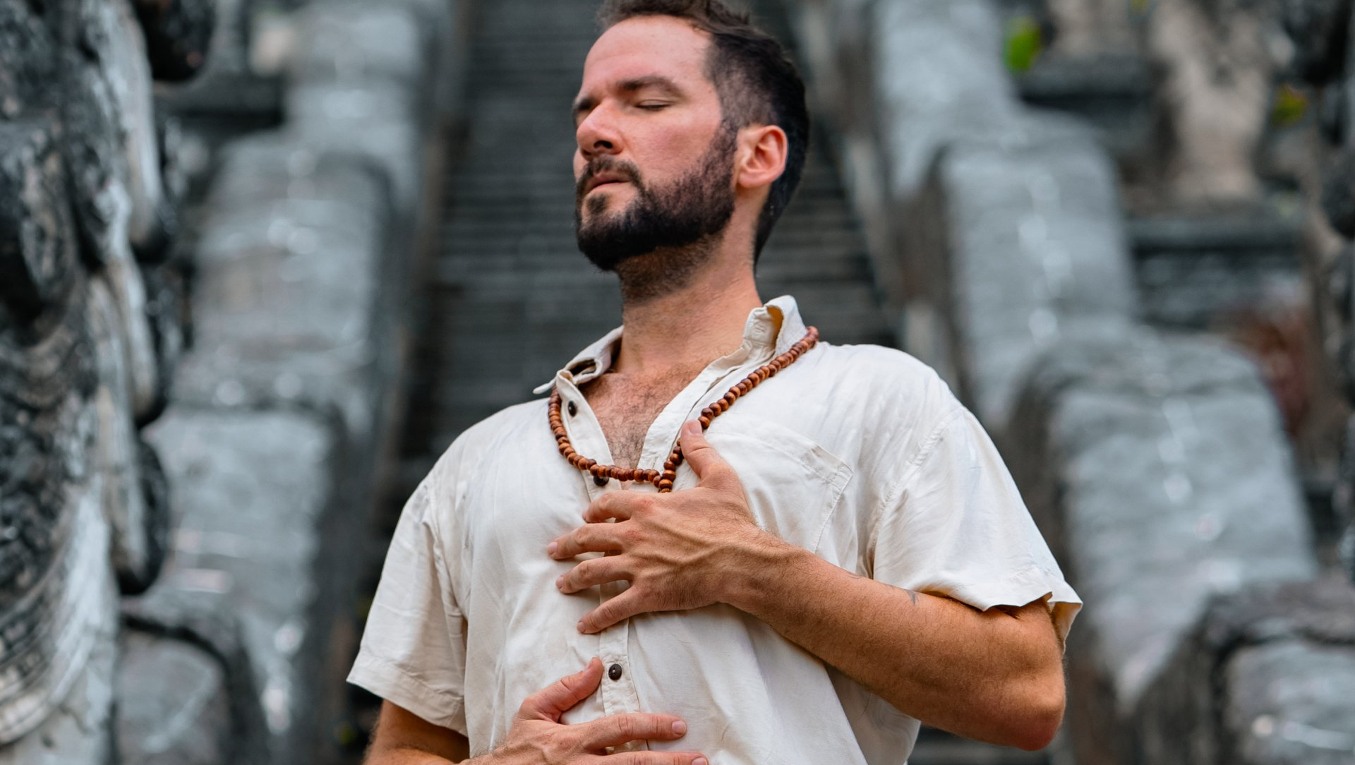 Man in light shirt meditating with eyes closed, hands on chest and stomach, on stone steps.