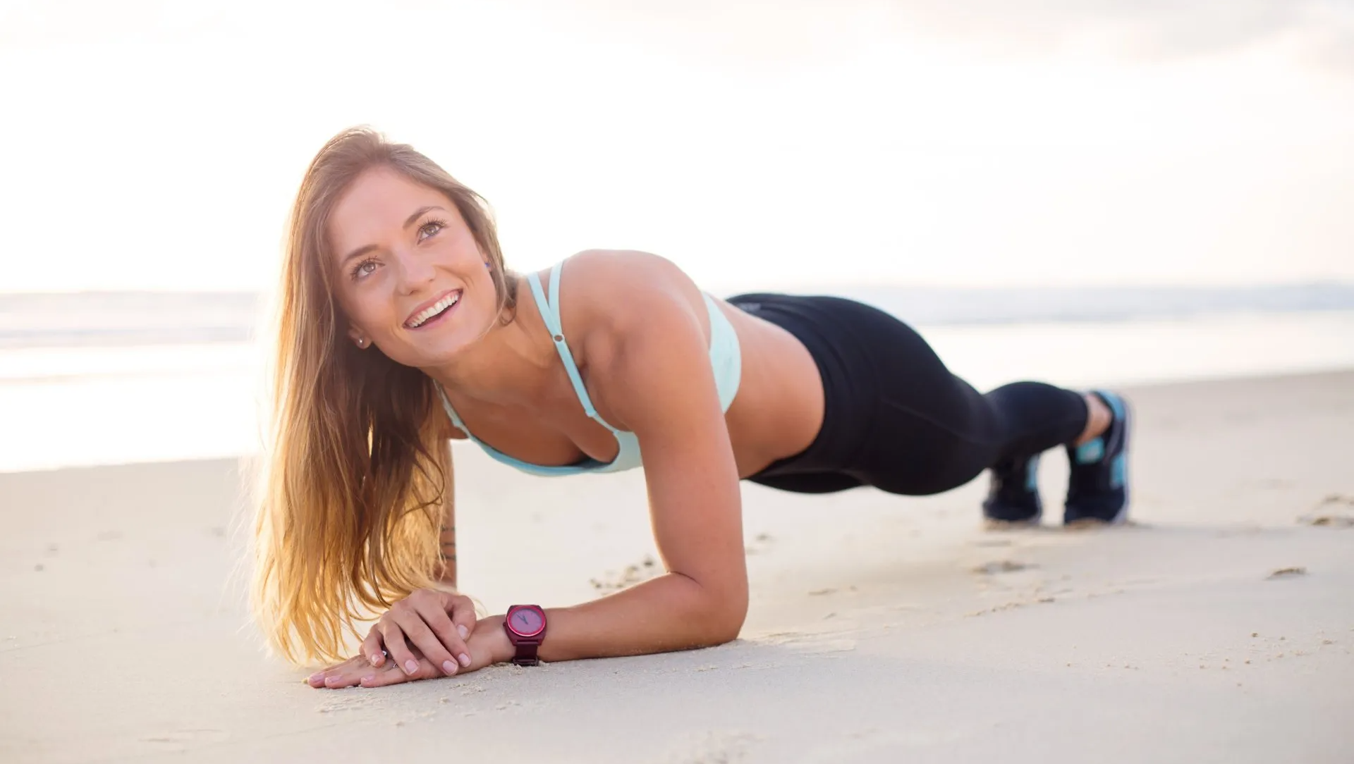 Woman doing plank exercise on a beach; wearing fitness attire, smiling.
