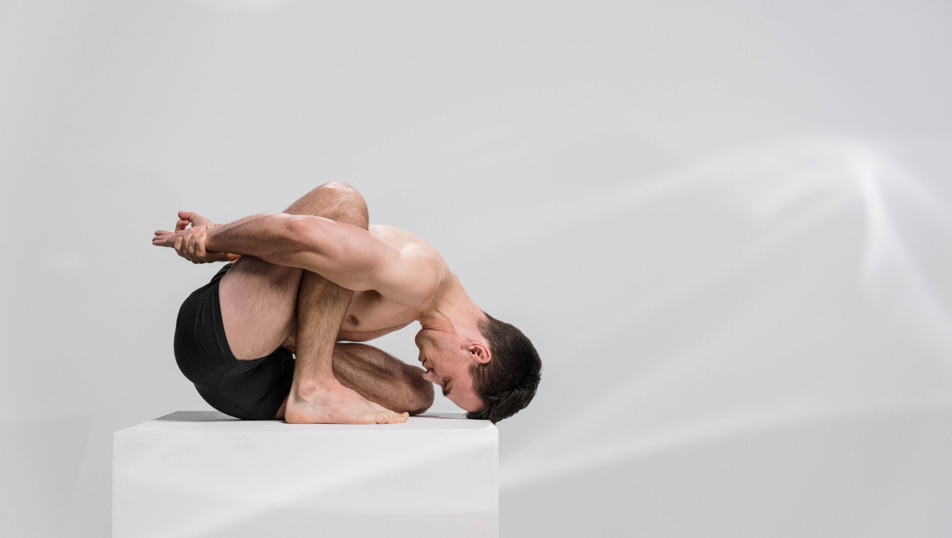 Man in yoga pose, crouched on a white block, arms behind back, head down.