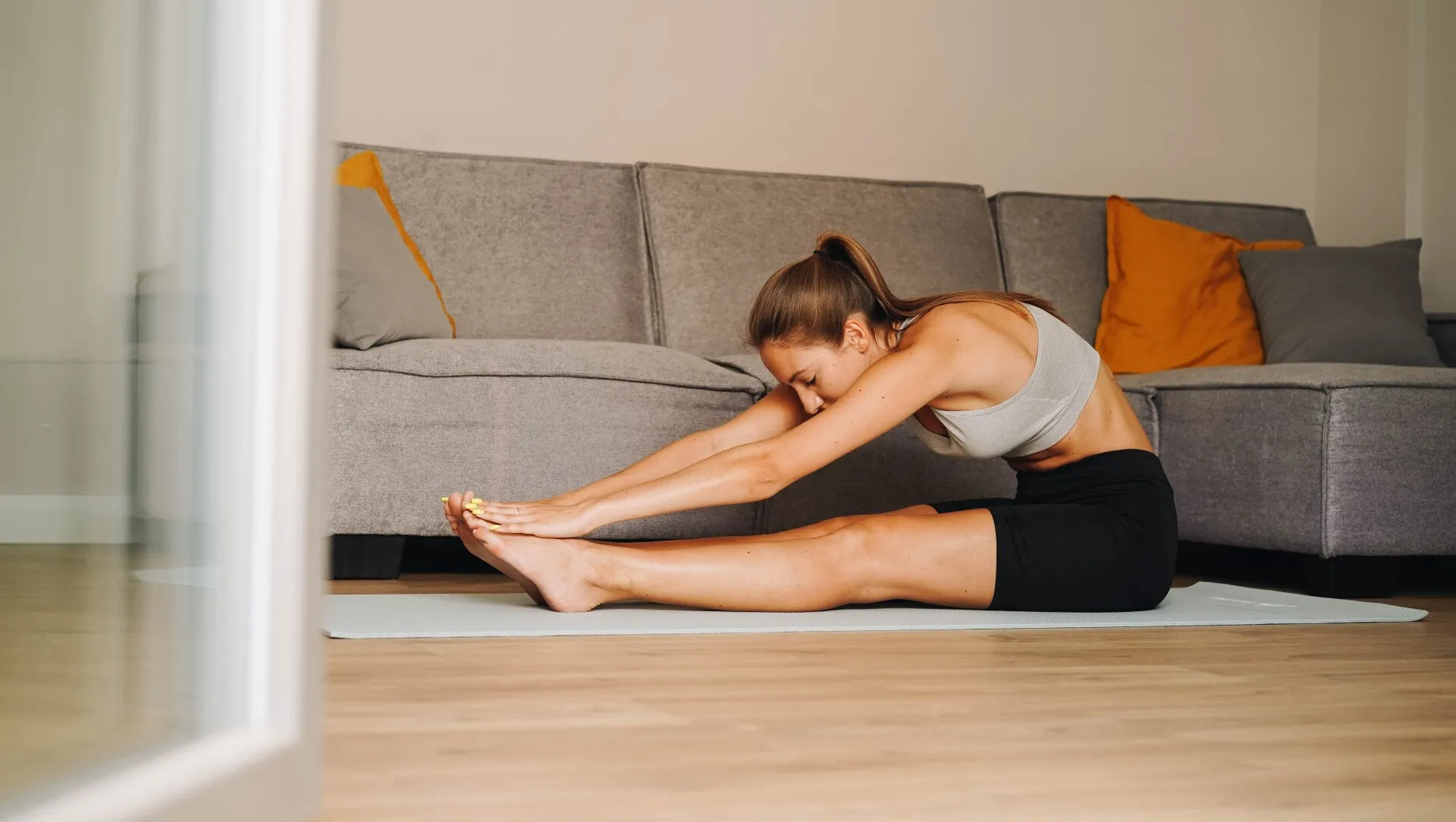 Woman stretching on a yoga mat in front of a gray couch, reaching for her toes.