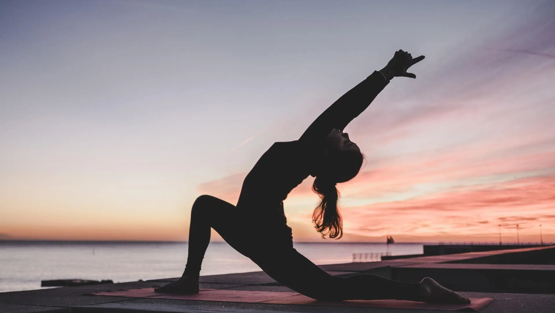 Silhouette of a person in a yoga pose with arm raised, ocean sunset backdrop.