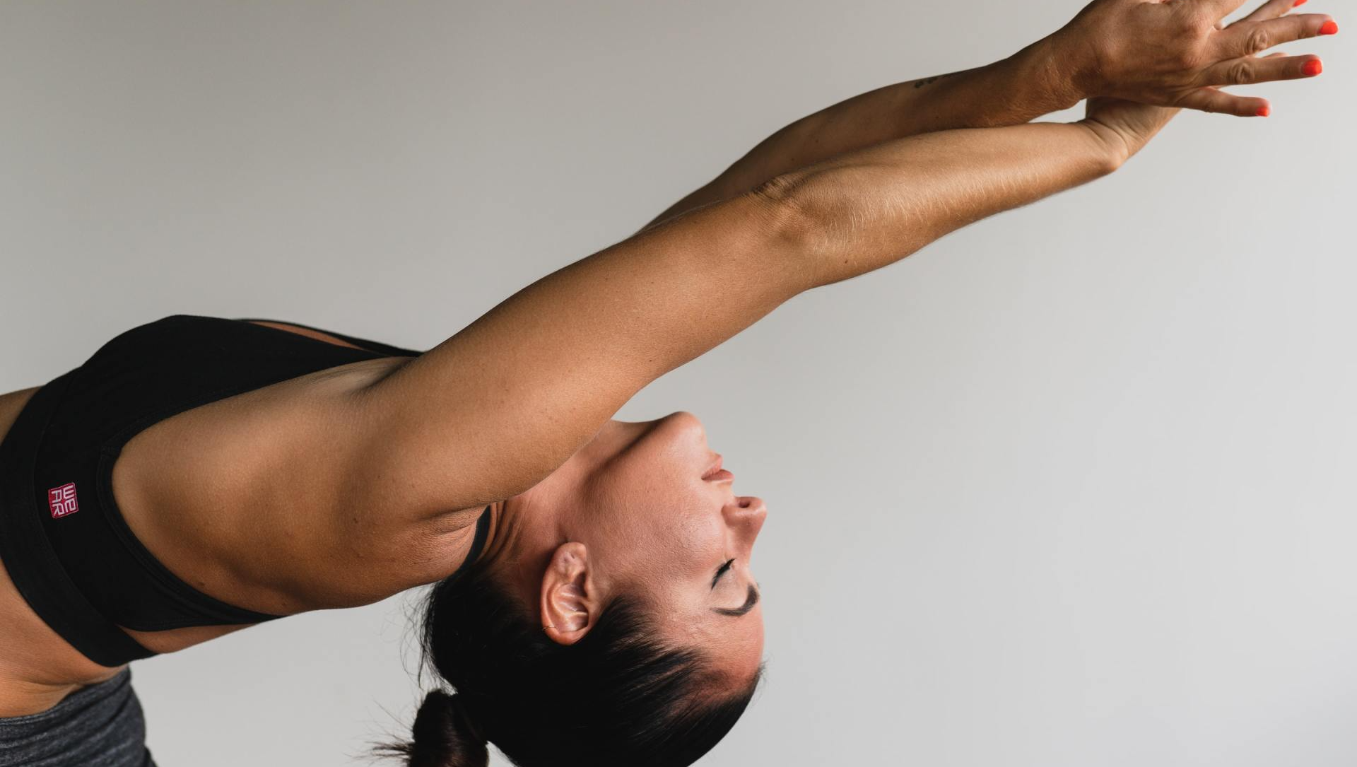 Woman in sports bra arches back, arms overhead, practicing yoga against a white background.