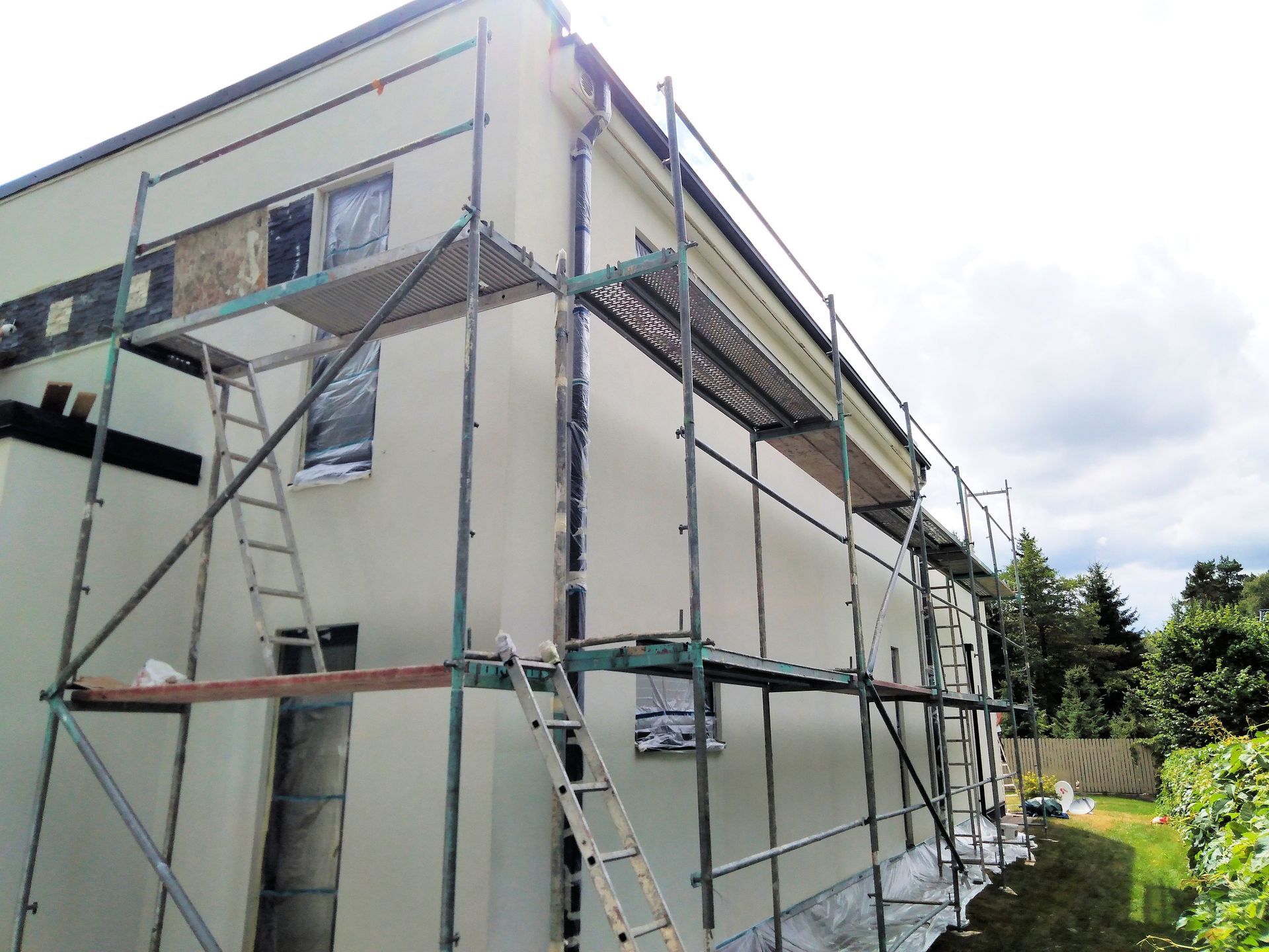 Scaffold around beige house under renovation with cloudy sky and garden beside it.