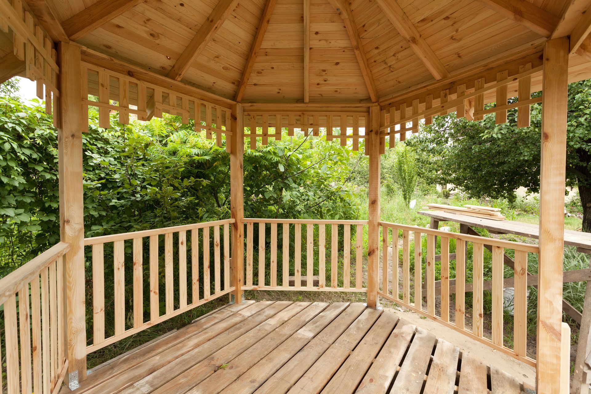 Wooden gazebo with railings and a peaked roof surrounded by lush green trees and a garden path.